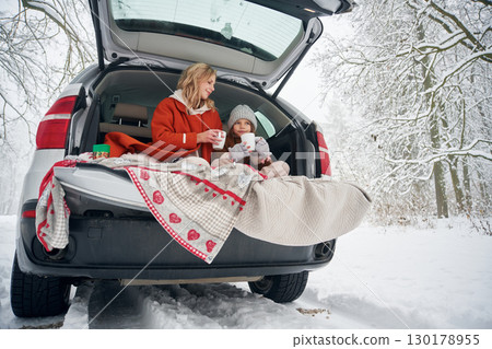 Drinking from the cup. Woman with her daughter is on the trunk of a car in the winter meadow and forest Drinking from the cup. Woman with her daughter is on the trunk of a car in the winter meadow and forest 130178955
