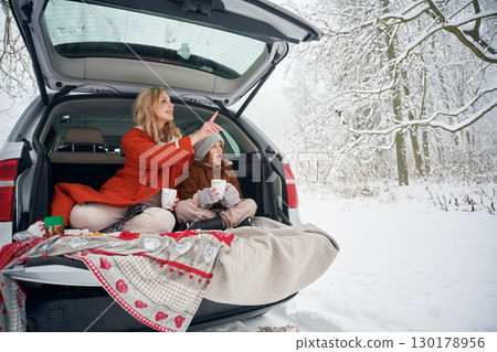 Drinking from the cup. Woman with her daughter is on the trunk of a car in the winter meadow and forest 130178956