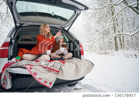 Drinking from the cup. Woman with her daughter is on the trunk of a car in the winter meadow and forest 130178957
