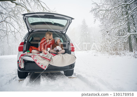 Woman with her daughter is on the trunk of a car in the winter meadow and forest Woman with her daughter is on the trunk of a car in the winter meadow and forest 130178958