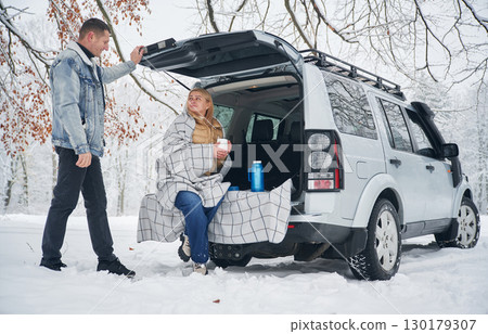 Woman is sitting in the trunk. Beautiful couple standing near the car in the winter forest Woman is sitting in the trunk. Beautiful couple standing near the car in the winter forest 130179307