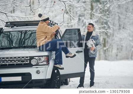 Woman is sitting on the car. Beautiful couple standing in the winter forest 130179308