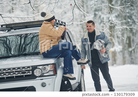 Woman is sitting on the car. Beautiful couple standing in the winter forest 130179309