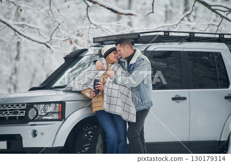 Man is covering woman with warm blanket. Beautiful couple standing near the car in the winter forest 130179314