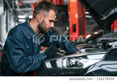 Busy car repairman is in the garage with automobile, fixing it Busy car repairman is in the garage with automobile, fixing it 130179458
