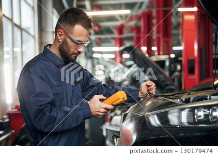 Professional electro device in hands. Car repairman is in the garage with automobile Professional electro device in hands. Car repairman is in the garage with automobile 130179474