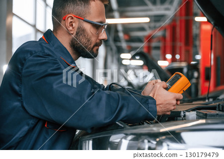 Yellow colored device for measuring electricity is in hands. Car repairman is in the garage with automobile Yellow colored device for measuring electricity is in hands. Car repairman is in the garage with automobile 130179479