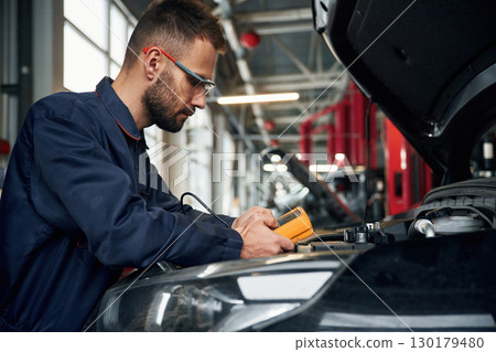 Yellow colored device for measuring electricity is in hands. Car repairman is in the garage with automobile 130179480