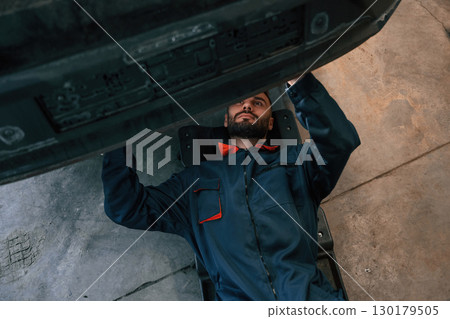 Lying down on the ground. Car repairman is in the garage with automobile 130179505