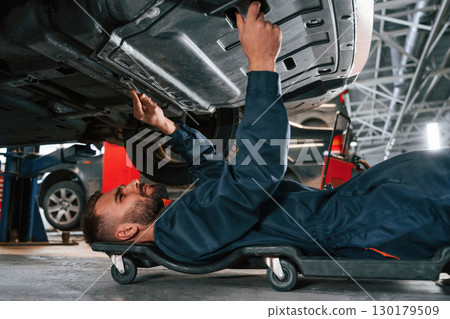Lying down on the ground. Car repairman is in the garage with automobile 130179509