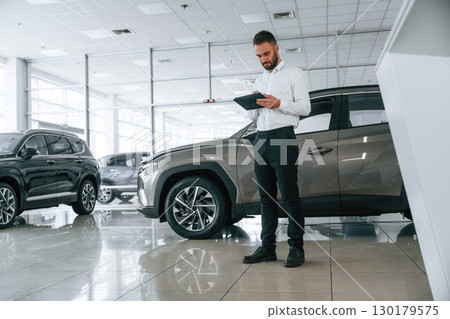 Black colored tablet in hands. Worker in formal clothes is in the auto salon Black colored tablet in hands. Worker in formal clothes is in the auto salon 130179575