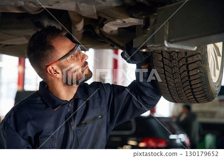 In protective eyewear, under vehicle. Car repairman is in the garage with automobile In protective eyewear, under vehicle. Car repairman is in the garage with automobile 130179652