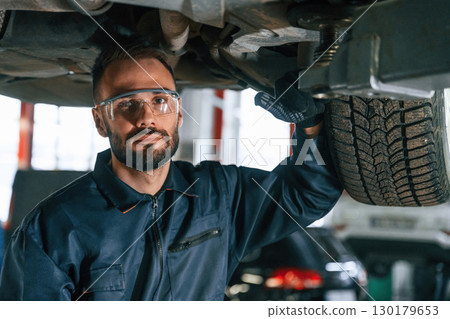 In protective eyewear, under vehicle. Car repairman is in the garage with automobile 130179653