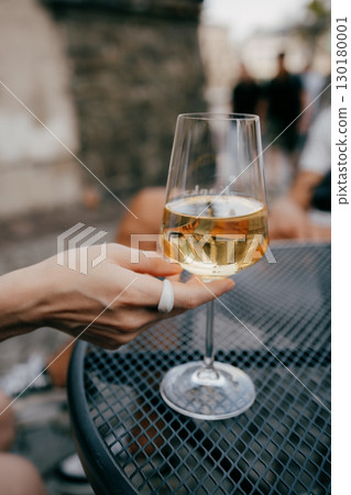 Woman with white ring holding glass of chilled white wine, sitting at metal table in an outdoor cafe, savoring tranquil moment under warm sunlight. Woman wearing white ring grasping white wine glass 130180001