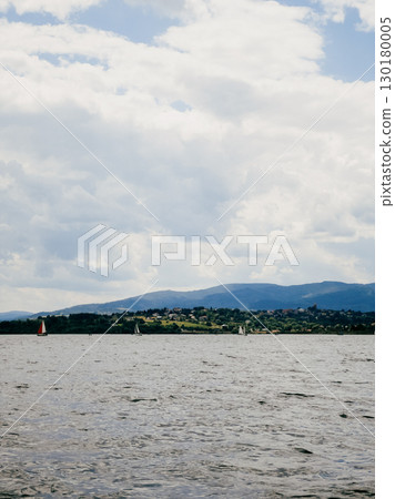 Sailboats are peacefully navigating lake on partly cloudy day, with mountains and small village visible in background, creating serene and picturesque scene. Sailboats gliding across calm lake waters 130180005