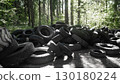 A large pile of discarded rubber tires is visible in a dense forest, showcasing the impact of pollution on the natural environment. 130180224