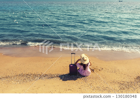 Young happy woman in pink dress and straw hat sitting near travel suitcase on the beach in summer. Woman holding straw hat. Summer, travel, and happy vacation concept Young happy woman in pink dress and straw hat sitting near travel suitcase on the beach in summer. Woman holding straw hat. Summer, travel, and happy vacation concept 130180348