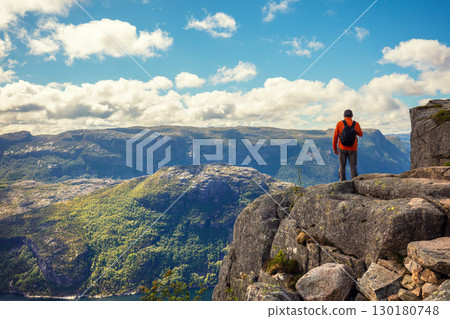 A man stands on a cliff edge. View of the beautiful Lysefjord on a cloudy day. Rocky shore of the majestic Lysefjord. Beautiful wild nature of Norway 130180748