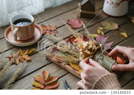 Woman is crafting an autumn decoration from dried leaves and flowers with cup of coffee nearby on rustic wooden table. Scene captures the warm cozy atmosphere of fall DIY project 130180835