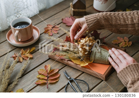 Cozy autumn scene with female hands arranging handmade decoration of dried flowers and leaves on wooden table, next to cup of coffee. Relaxing and creative fall activity 130180836