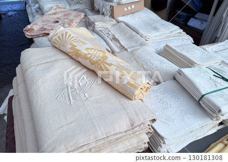 Stacks of embroidered and patterned vintage linens displayed on a table at a flea market. Flea market, textile preservation, monogrammed fabrics, heritage embroidery Stacks of embroidered and patterned vintage linens displayed on a table at a flea market. Flea market, textile preservation, monogrammed fabrics, heritage embroidery 130181308