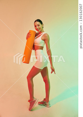 Orange colored yoga mat in hands. Young fitness woman in sportive clothes against background in studio 130182007
