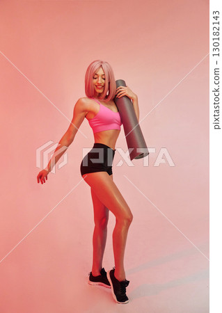 Holding yoga mat. Sportive woman with pink wig is against background Holding yoga mat. Sportive woman with pink wig is against background 130182143