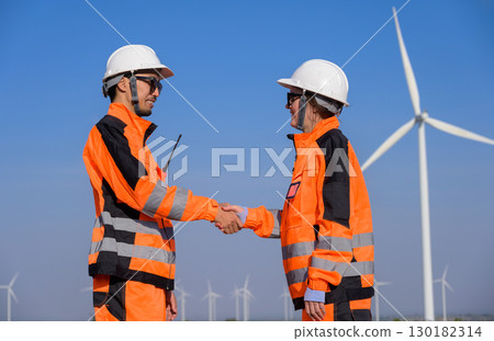Two workers in orange uniform shaking hands while standing in wind turbine farm 130182314