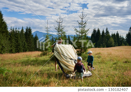 Man hiker and two young boys work together to set up tourist tent on grassy hillside. Pine trees and distant mountains under partly cloudy sky. 130182407