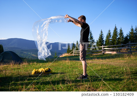 Man hiker set up tourist tent on grassy hillside. Pine trees and distant mountains under clear blue sky. Man hiker set up tourist tent on grassy hillside. Pine trees and distant mountains under clear blue sky. 130182409