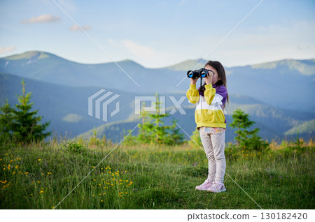 Young girl stands in grassy meadow, looking through binoculars, wearing yellow and purple sweatshirt. Behind her, rolling green hills and mountains stretch into distance under clear blue sky. Young girl stands in grassy meadow, looking through binoculars, wearing yellow and purple sweatshirt. Behind her, rolling green hills and mountains stretch into distance under clear blue sky. 130182420
