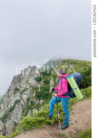 Woman hiker stands on misty mountain trail, gripping trekking poles. Female tourist with green sleeping pad strapped backpack and gazes at rugged, rocky cliffs partially shrouded in fog. 130182422