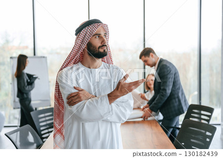 Standing in front of people that behind. Muslim businessman in traditional outfit with colleagues in office Standing in front of people that behind. Muslim businessman in traditional outfit with colleagues in office 130182439