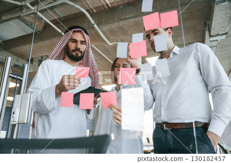 Using stickers on the glass. Muslim businessman in traditional outfit with colleagues in office Using stickers on the glass. Muslim businessman in traditional outfit with colleagues in office 130182457