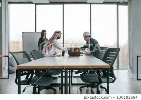 Working with a plan. Muslim businessman in traditional outfit with colleagues in office Working with a plan. Muslim businessman in traditional outfit with colleagues in office 130182684