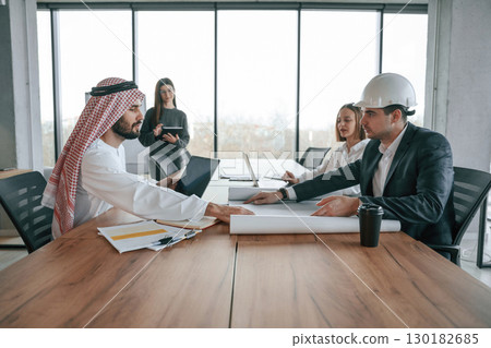 Working with a plan. Muslim businessman in traditional outfit with colleagues in office Working with a plan. Muslim businessman in traditional outfit with colleagues in office 130182685
