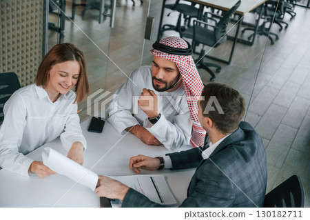 Sitting by the table. Muslim businessman in traditional outfit with colleagues in office Sitting by the table. Muslim businessman in traditional outfit with colleagues in office 130182711