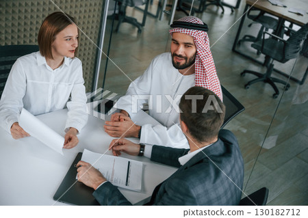 Sitting by the table. Muslim businessman in traditional outfit with colleagues in office Sitting by the table. Muslim businessman in traditional outfit with colleagues in office 130182712