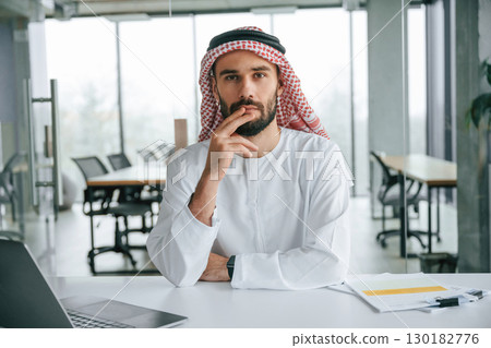 By the table with laptop. Successful Muslim businessman in traditional outfit in his office By the table with laptop. Successful Muslim businessman in traditional outfit in his office 130182776