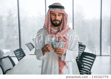 Holding money in hands. Successful Muslim businessman in traditional outfit in his office Holding money in hands. Successful Muslim businessman in traditional outfit in his office 130182779