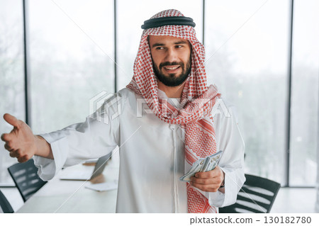 Holding money in hands. Successful Muslim businessman in traditional outfit in his office 130182780