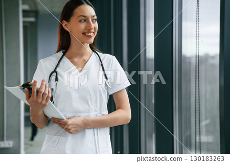 Standing and holding documents in hands. Female doctor in white coat is indoors Standing and holding documents in hands. Female doctor in white coat is indoors 130183263