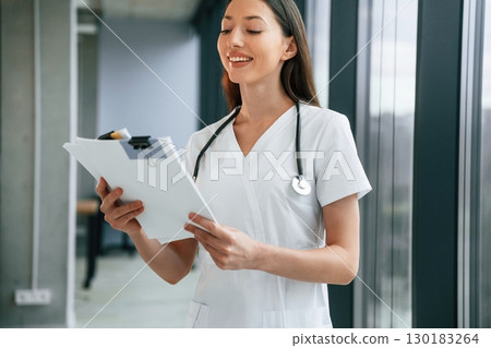 Standing and holding documents in hands. Female doctor in white coat is indoors 130183264