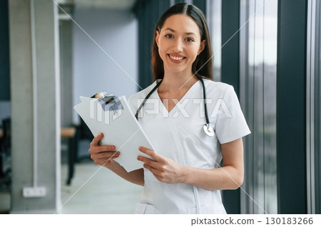 Holding notepad in hands. Female doctor in white coat is indoors 130183266
