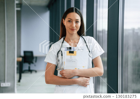 Holding notepad in hands. Female doctor in white coat is indoors 130183275