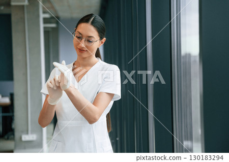 Preparing for work, wearing gloves. Female doctor in white coat is indoors 130183294