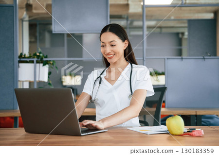 Nutritionist with apple and laptop. Female doctor in white coat is indoors Nutritionist with apple and laptop. Female doctor in white coat is indoors 130183359