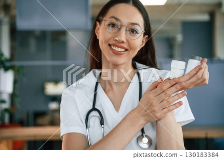 In glasses. Sitting by the table. Female doctor in white coat is indoors In glasses. Sitting by the table. Female doctor in white coat is indoors 130183384