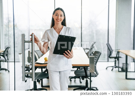 Front view. Standing with notepad in hands. Female doctor in white coat is indoors 130183393