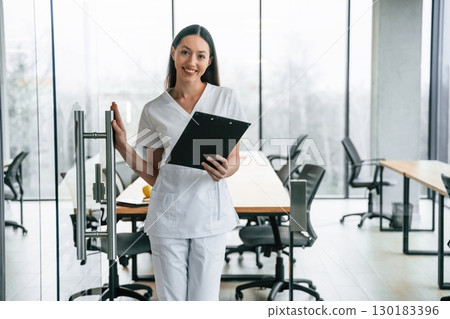 Front view. Standing with notepad in hands. Female doctor in white coat is indoors 130183396